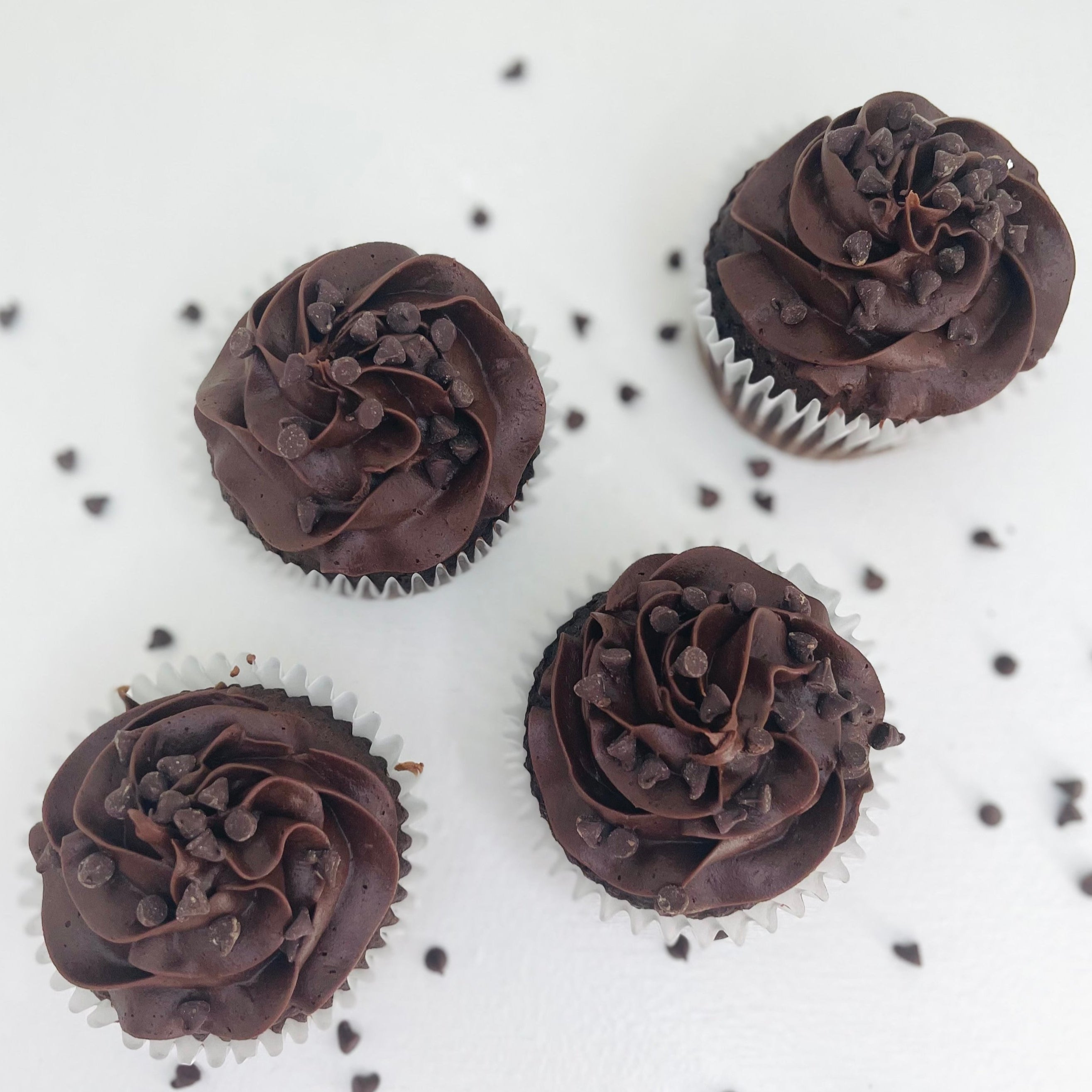 A set of four jumbo chocolate cupcakes with chocolate buttercream frosting and chocolate shavings on top, displayed on a white surface with scattered chocolate chips.