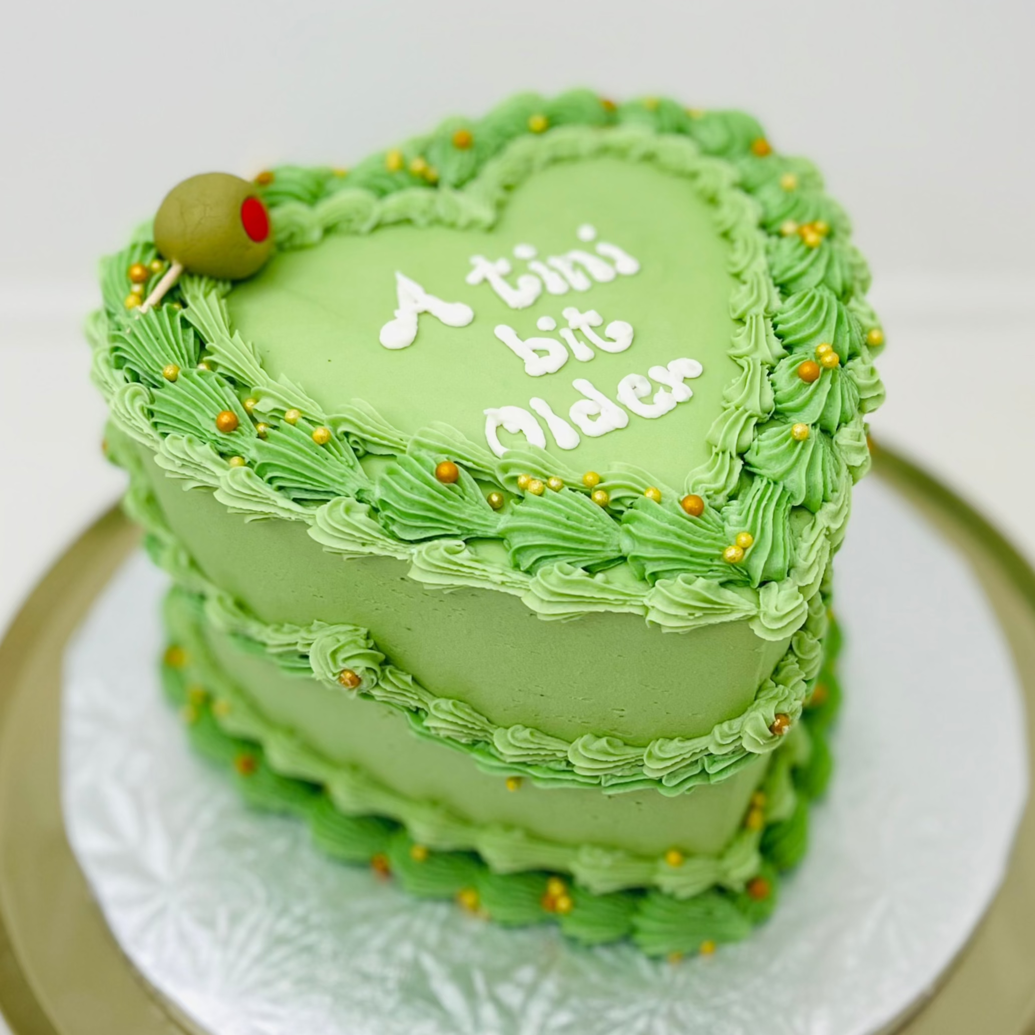 A green, heart-shaped cake with vintage buttercream frosting and a message 'A tini bit older' on top.