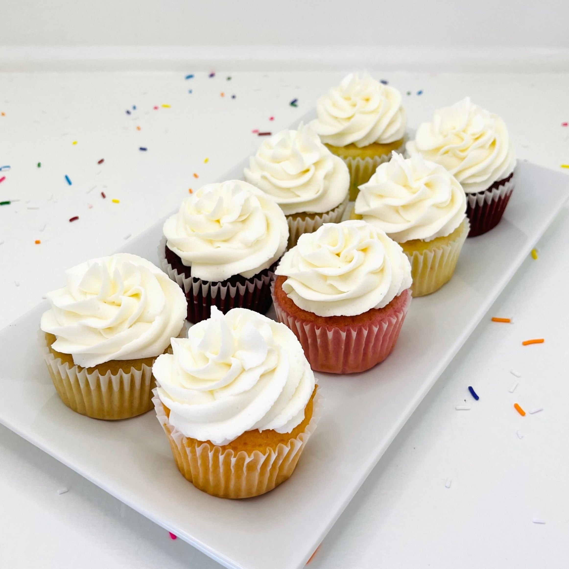 Assorted cupcakes with white frosting on a white plate with colorful sprinkles.