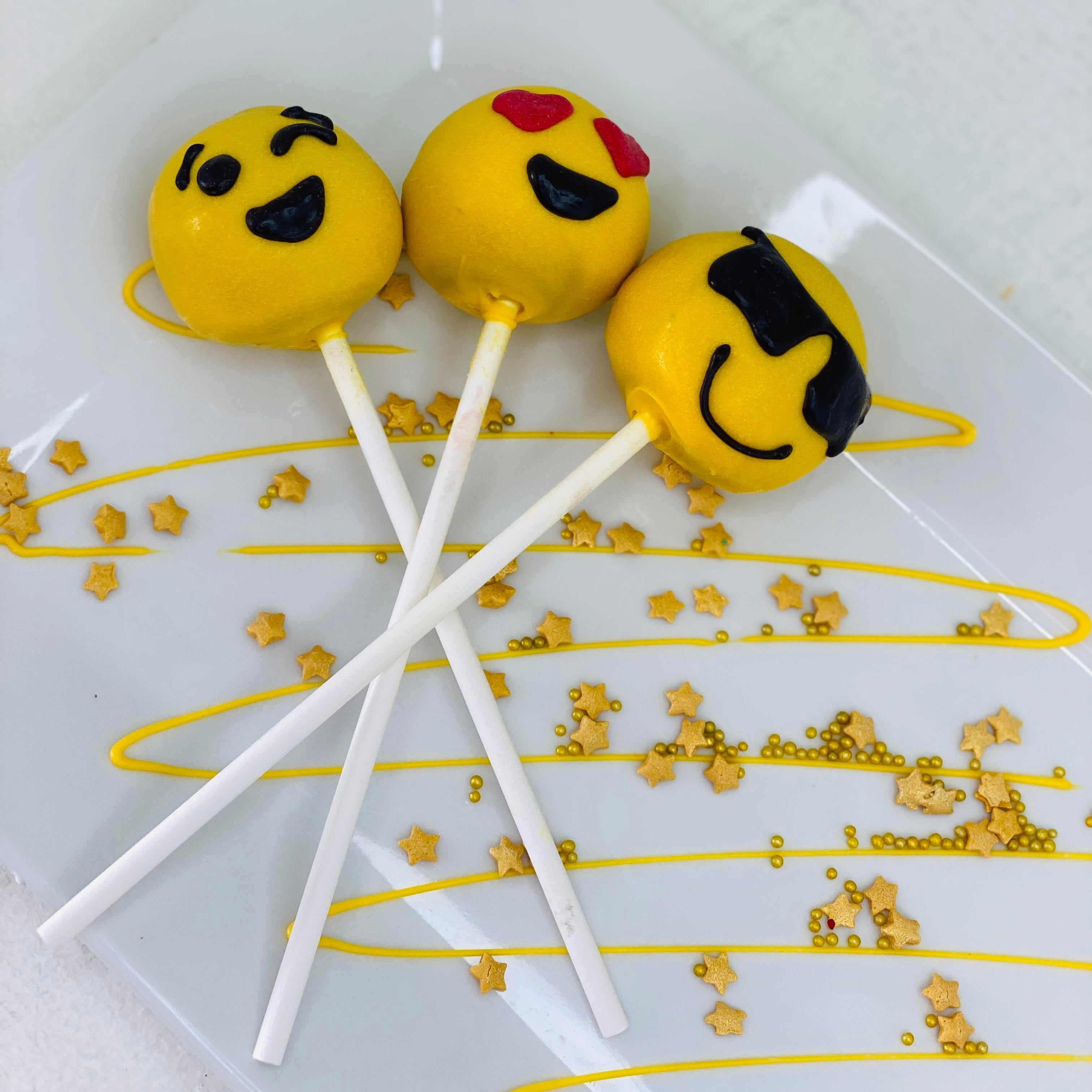 Three emoji-themed cake pops on a decorative plate with gold accents.