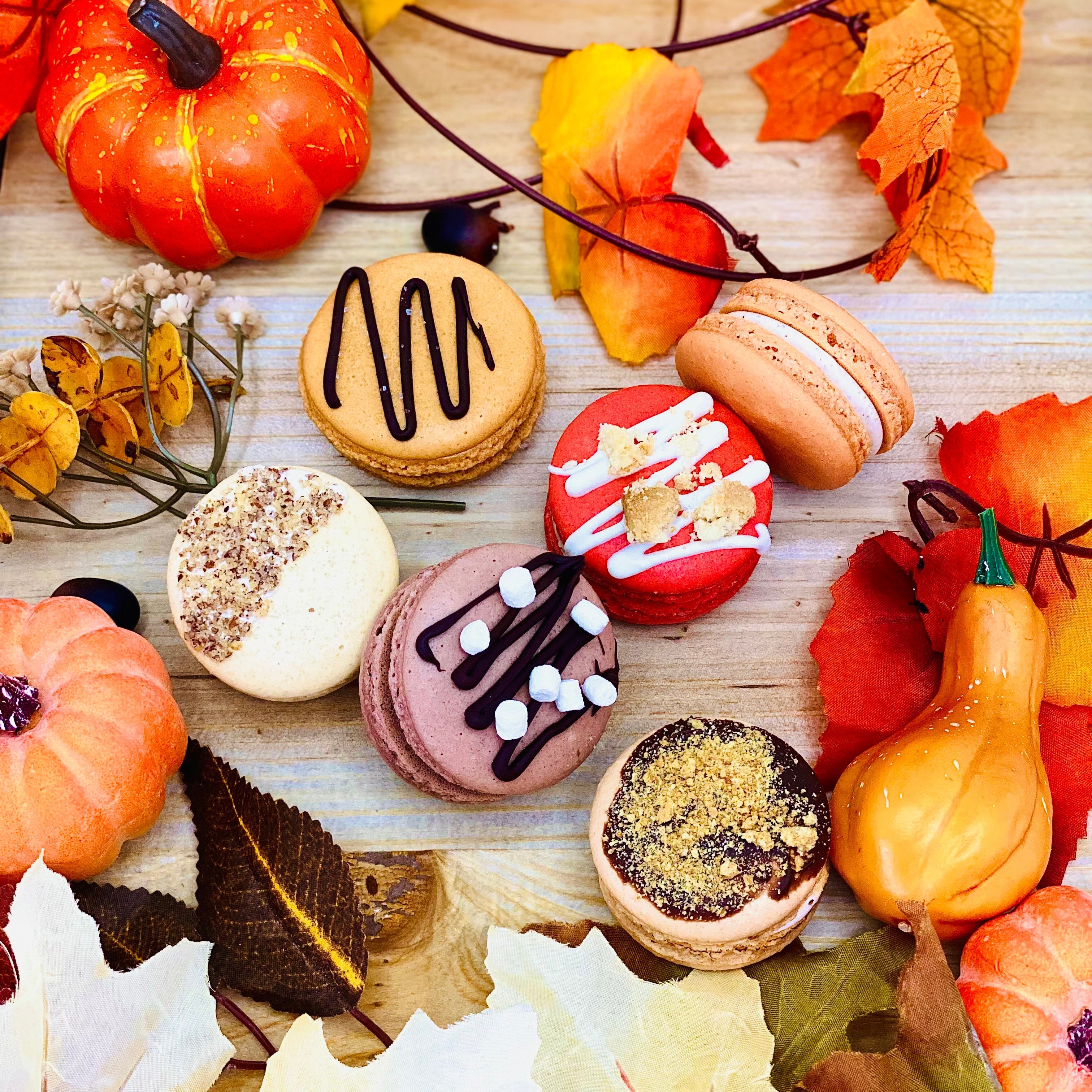 Assorted macarons with autumn decorations including pumpkins and leaves on a wooden surface.