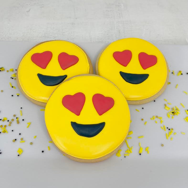 Three cookies shaped like smiling emojis with heart eyes on a white surface.