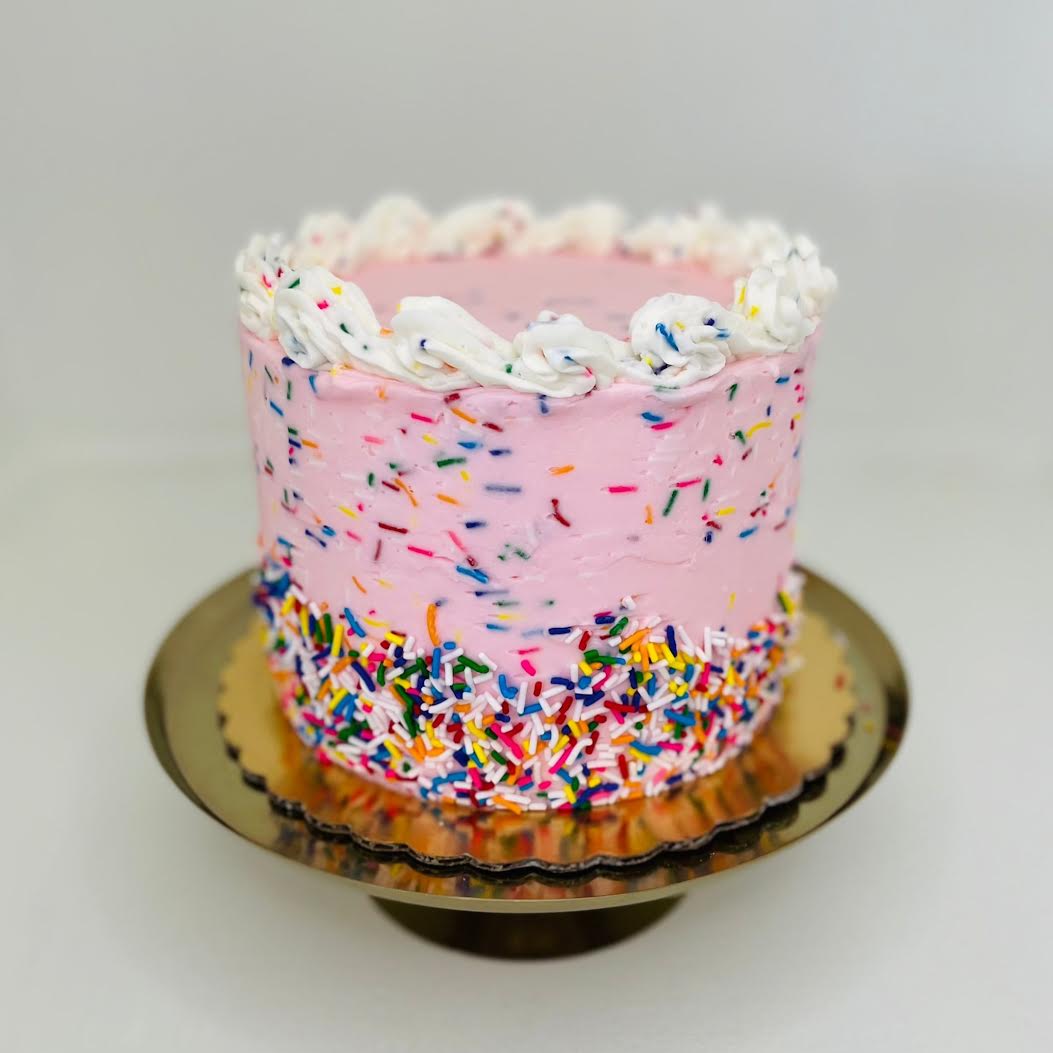 Pink cake with colorful sprinkles on a gold cake stand against a light gray background