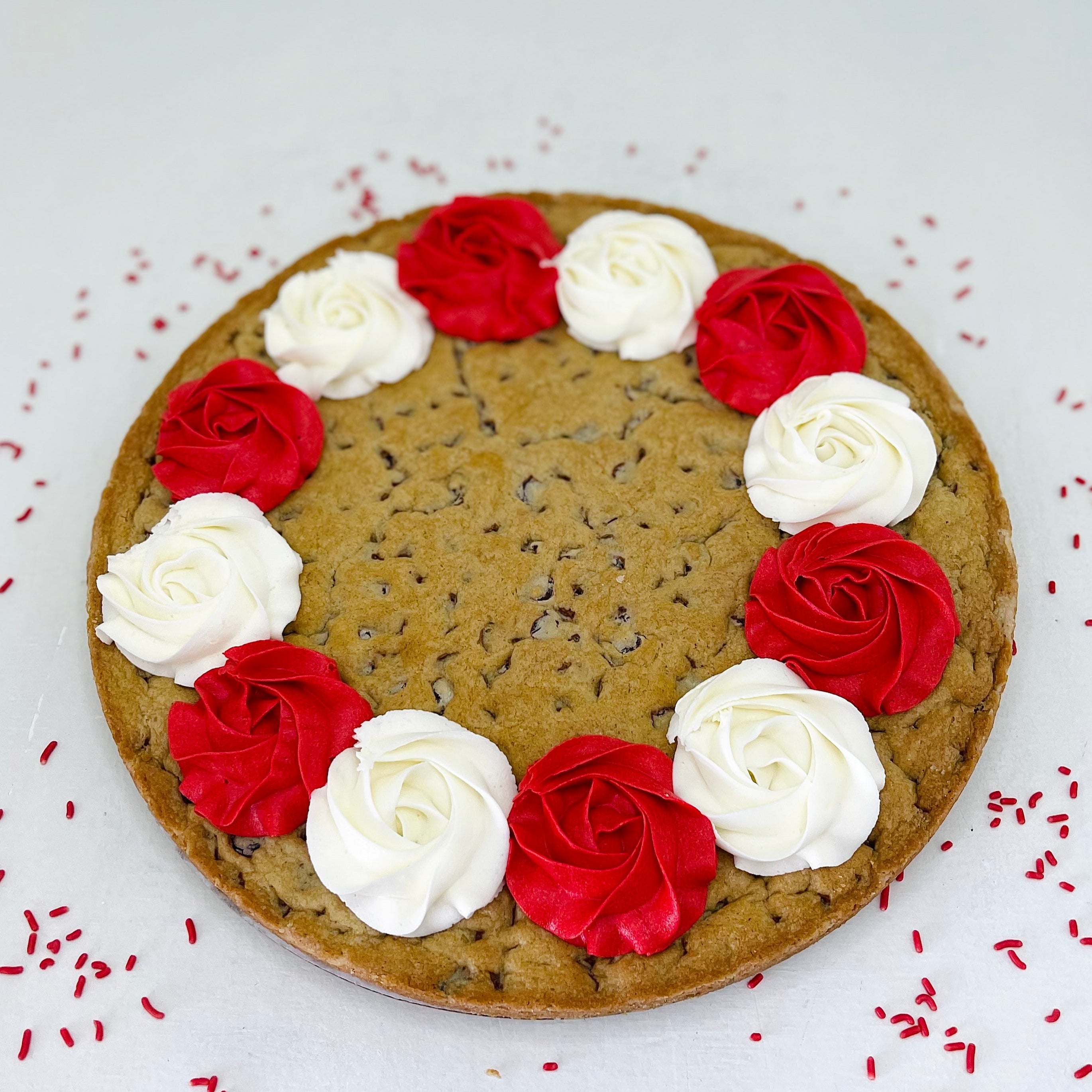 Large cookie decorated with red and white rose-shaped candies on a white background with red sprinkles.