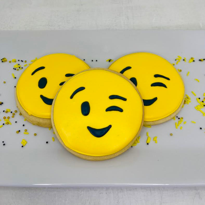 Three yellow smiley face wink cookies with black accents on a white tray.